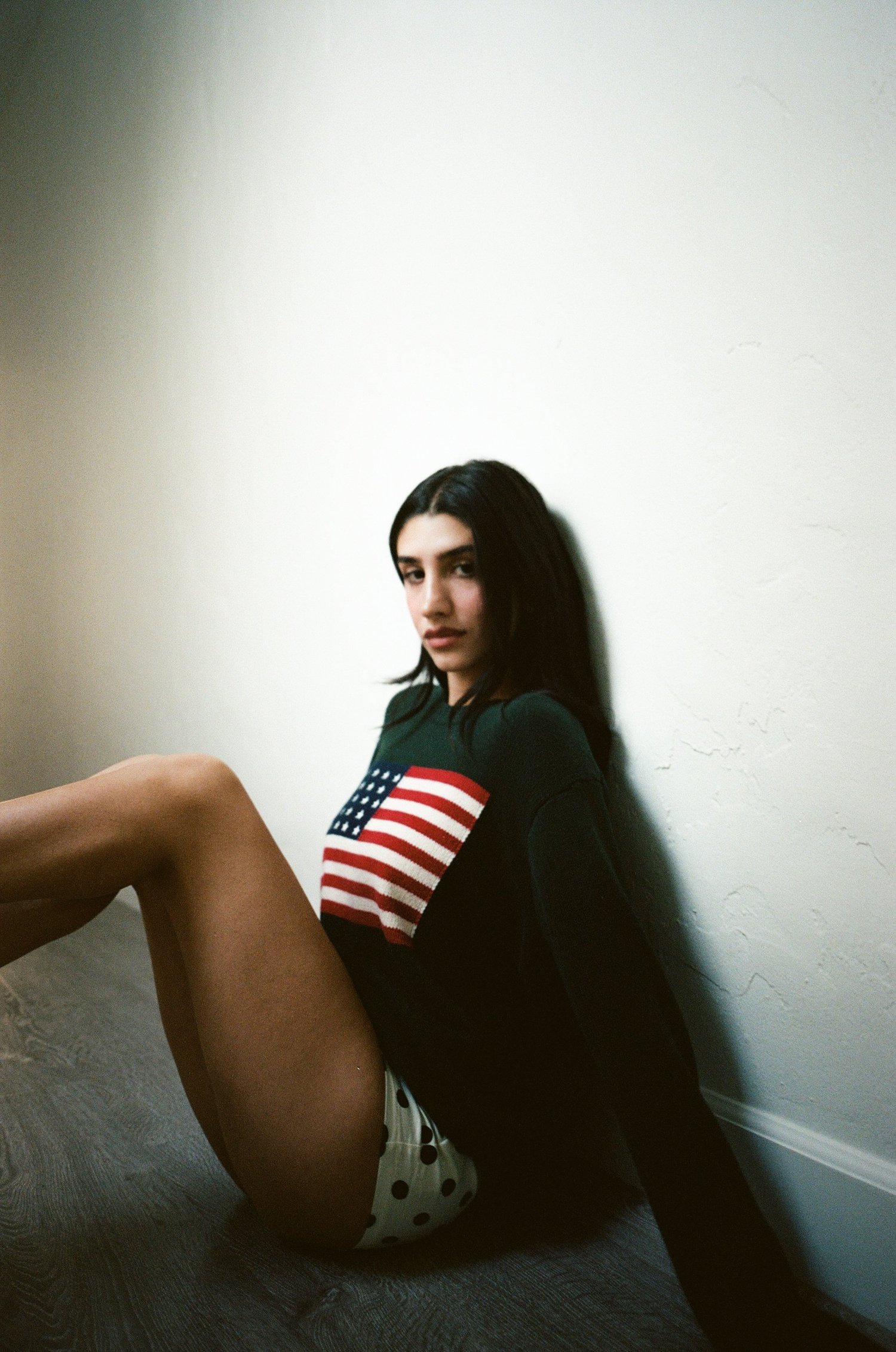 Portrait of Kayel seated against a white wall wearing a flag sweater