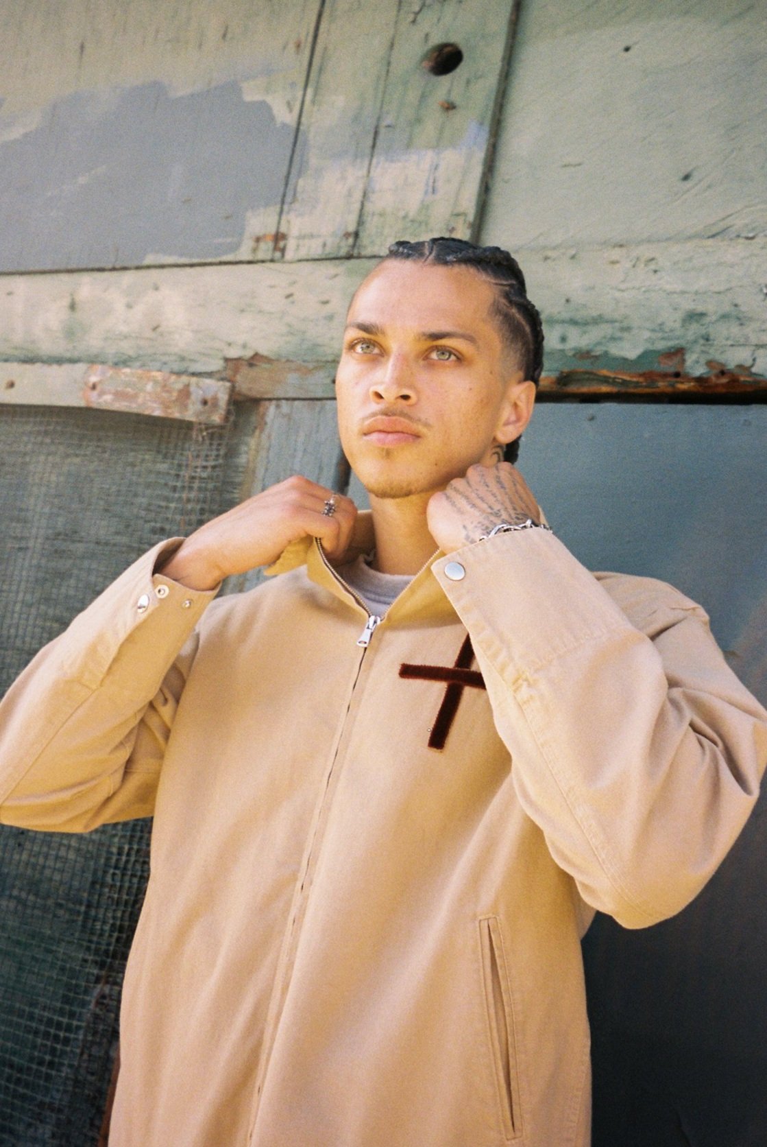 Model in a sand-coloured jacket against green corrugated metal — Trinity Drop 2